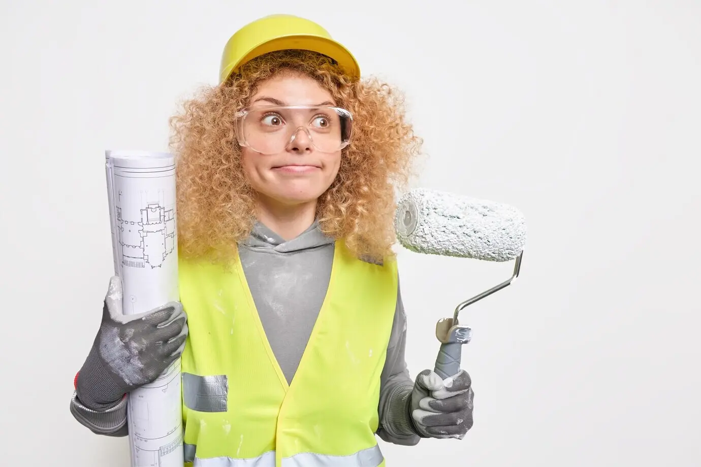Eine Frau mit zusammengepressten Lippen blickt konzentriert in die Ferne, trägt Schutzhelm, Uniform und Schutzbrille; als professionelle Dekorateurin hält sie eine Rolle und einen Bauplan und posiert drinnen. Heimreparaturservice.